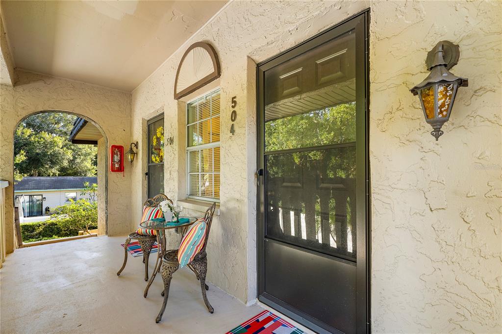 A covered porch features a small bistro set, a screen door, and a wall-mounted lamp — 504 CORDOVA GREEN, SEMINOLE, FL