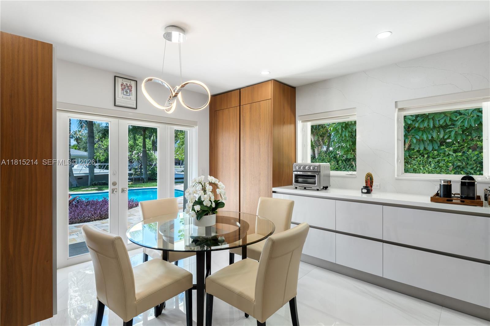 A modern dining area features a glass table, cream chairs, wood cabinetry, and pool views — 435 Marquesa Dr, Coral Gables FL 33156, CORAL GABLES, FL