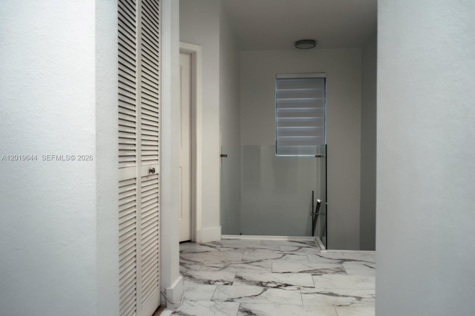 A hallway with white walls, marble-look tile flooring, a louvered closet door, and glass railing — 4180 NE 21st St, Homestead FL 33033, HOMESTEAD, FL