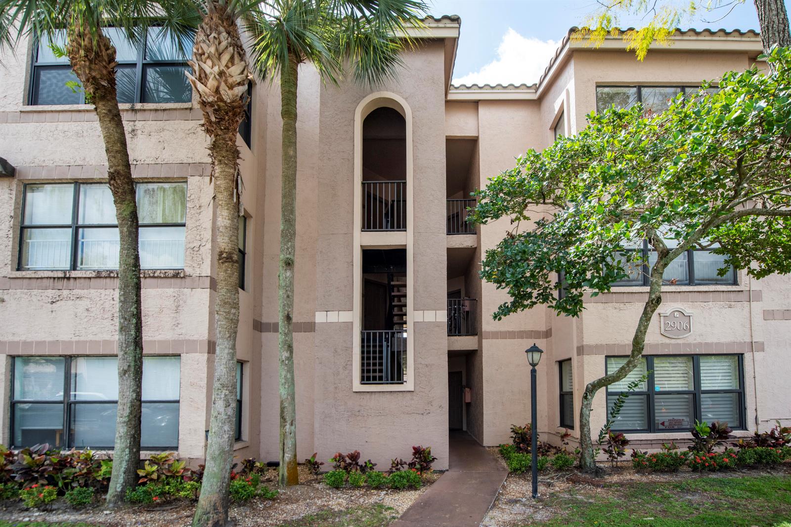 A three-story tan stucco apartment building features a central arched stairwell and palm trees — 2900 S University Drive # 9210, Davie FL 33328, DAVIE, FL