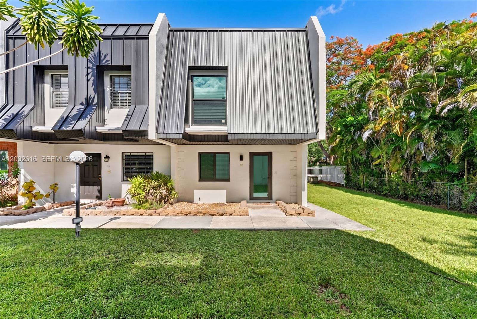 A two-story townhouse features a metal roof, white stucco walls, and a grassy front yard — 5555 Ludlam Rd # 23, South Miami FL 33155, SOUTH MIAMI, FL
