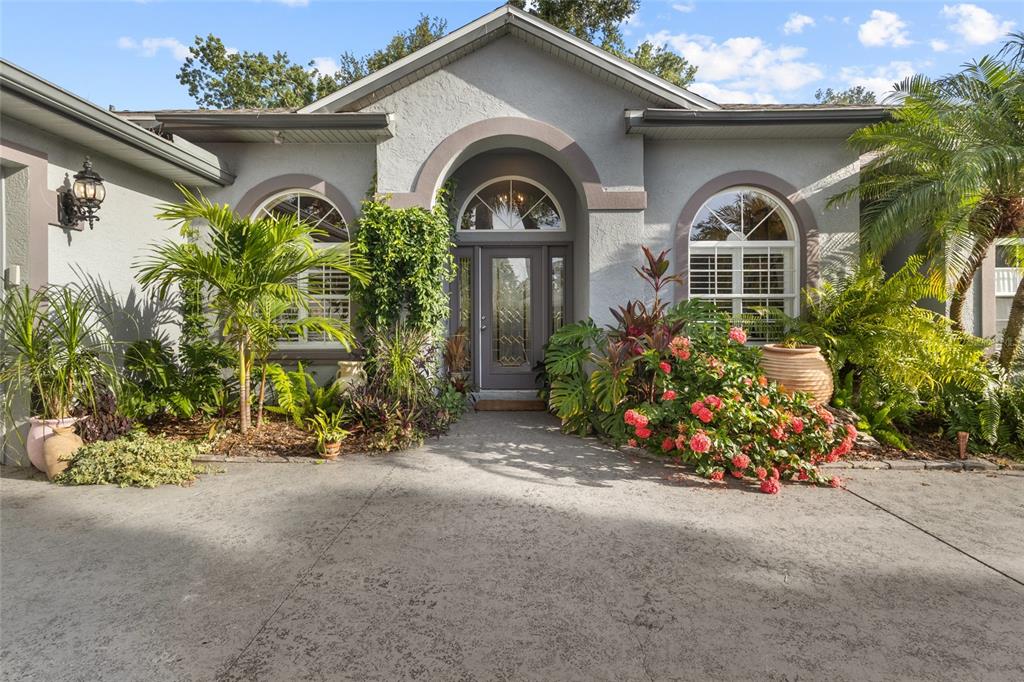 A gray stucco house features an arched entryway, a glass-paneled front door, and lush landscaping — 3025 RIVER WOODS DRIVE, PARRISH, FL