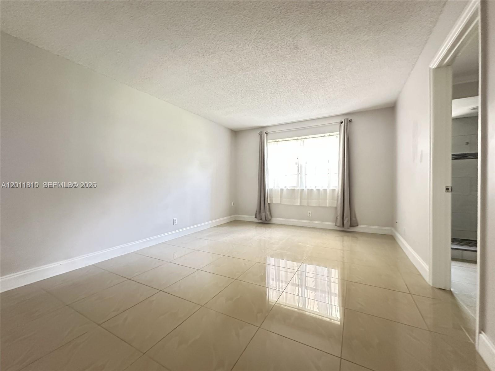 An empty room features light-colored tile flooring, white walls, a window with curtains, and a doorway — 9370 SW 8th St # 107, Boca Raton FL 33428, BOCA RATON, FL