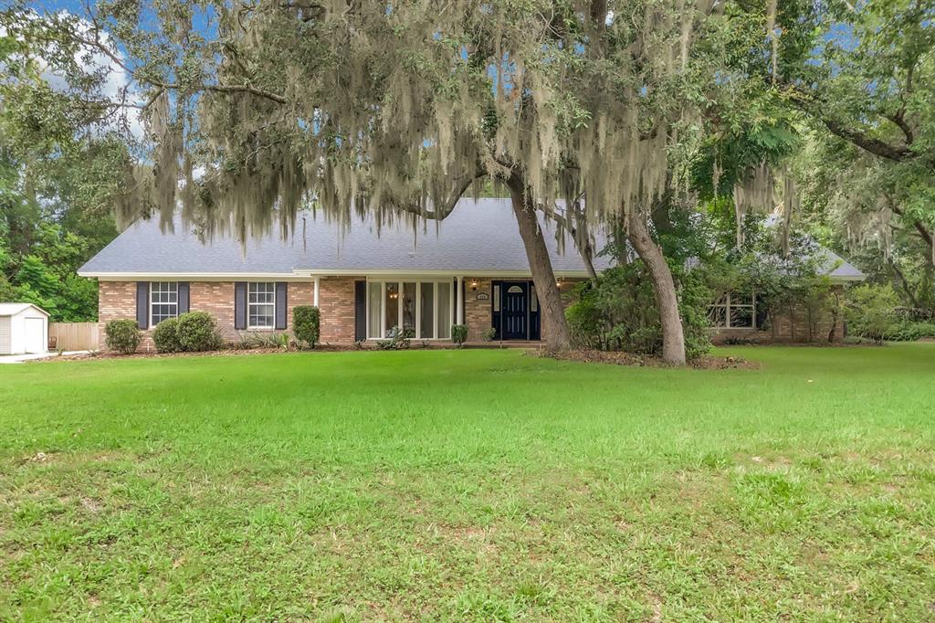 A single-story brick house with a dark roof sits behind a large lawn and trees — 112 FOREST PARK COURT, LONGWOOD, FL