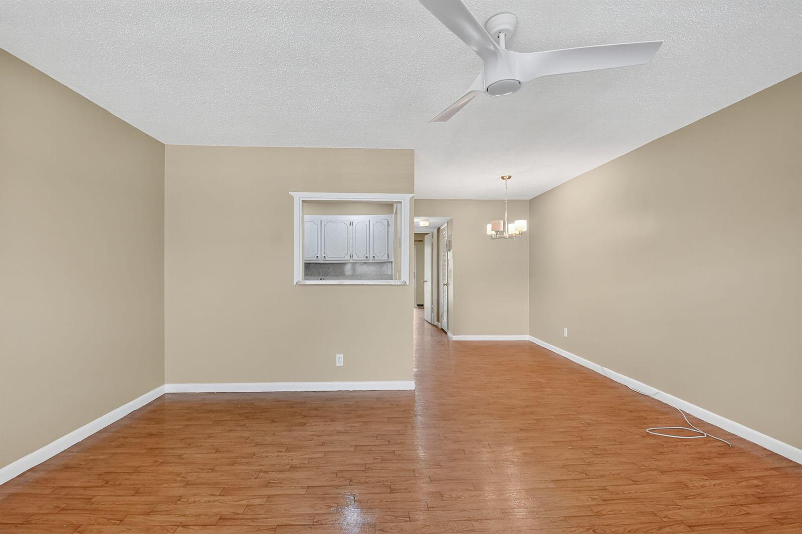 An empty living room with beige walls, wood-look flooring, a ceiling fan, and a pass-through window — 265 Windsor # L, West Palm Beach FL 33417, WEST PALM BEACH, FL