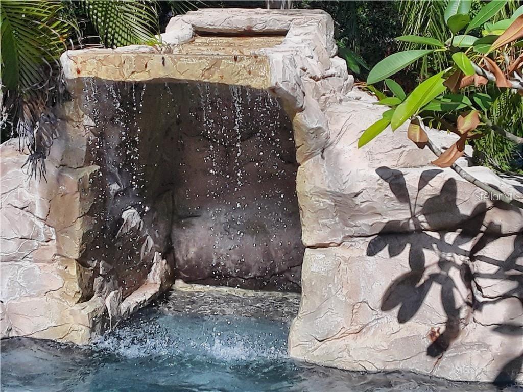 A stone-textured pool waterfall feature cascades into a swimming pool surrounded by tropical plants — 681 JASMINE WAY S, ST PETERSBURG, FL
