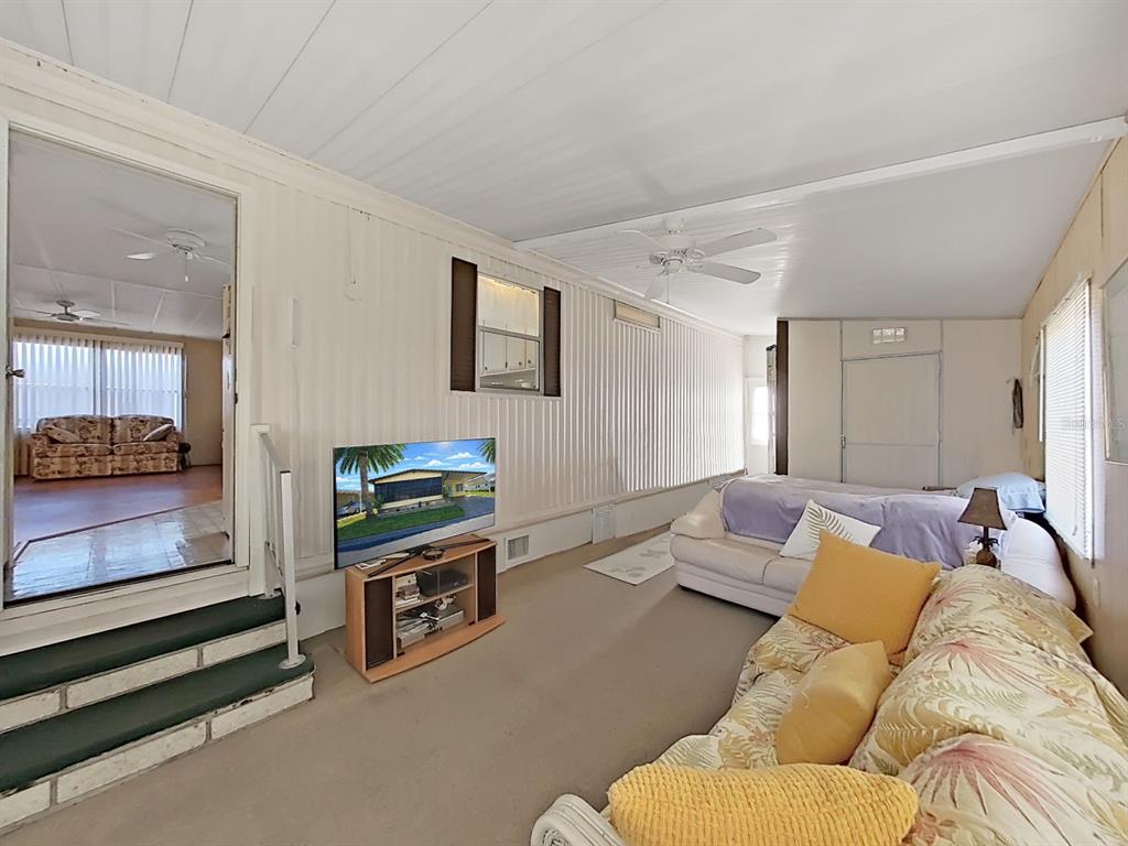 A living room with carpeted floors, a television, and a view into an adjacent sunroom — 12501 ULMERTON ROAD UNIT 140, LARGO, FL