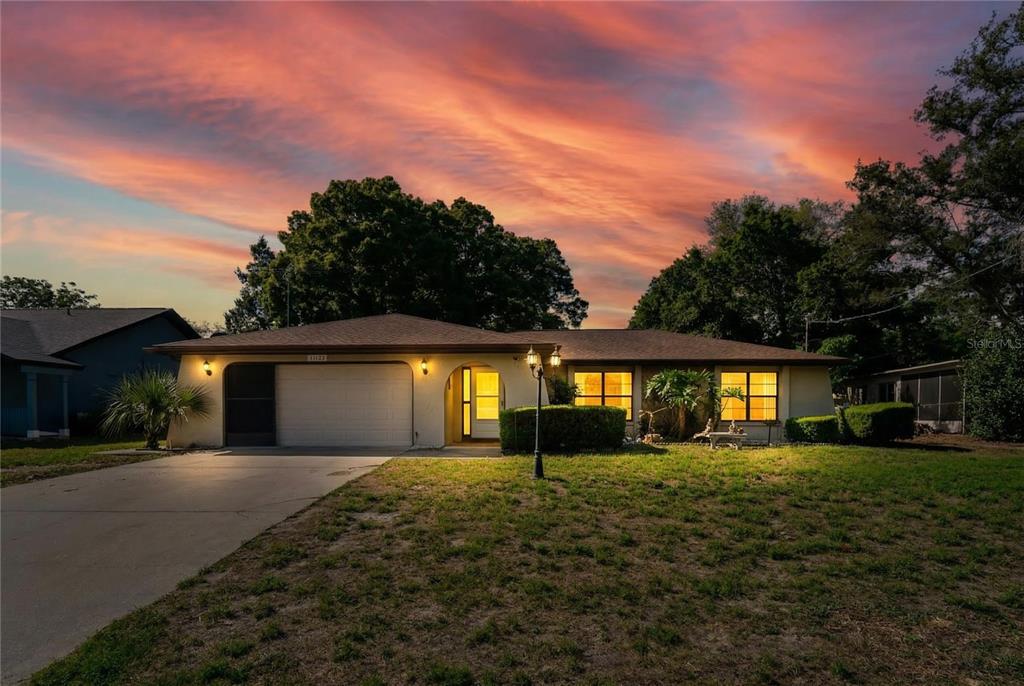 A single-story house with a two-car garage and a sunset sky in the background — 11122 MADERIA STREET, SPRING HILL, FL
