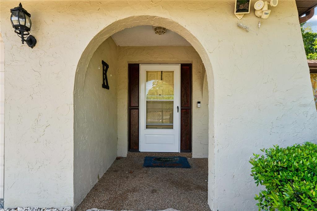 A covered entryway features a white storm door, arched stucco walls, and a textured floor — 11122 MADERIA STREET, SPRING HILL, FL