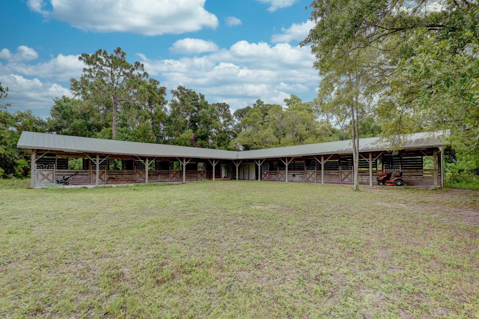 A long, L-shaped wooden horse stable with a metal roof sits in a grassy field — 19153 Eagles Way Court, Loxahatchee FL 33470, LOXAHATCHEE, FL