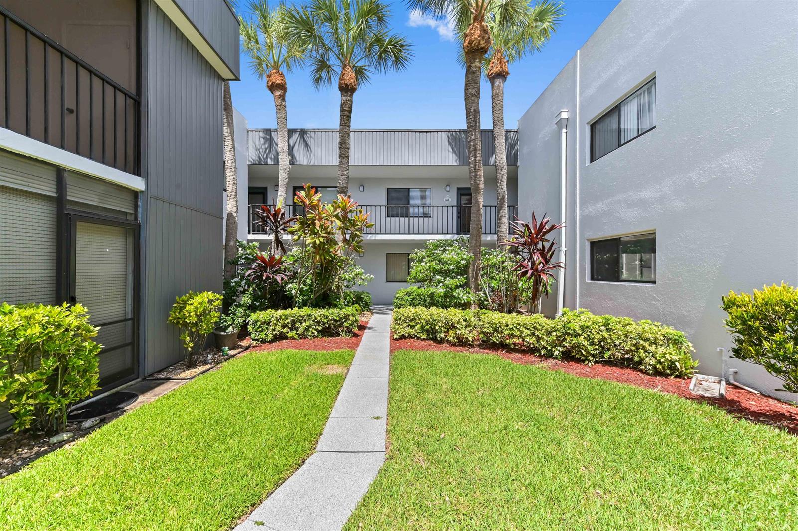 A concrete walkway leads through a grassy courtyard between two-story apartment buildings with palm trees — 15054 Ashland Way # 84, Delray Beach FL 33484, DELRAY BEACH, FL