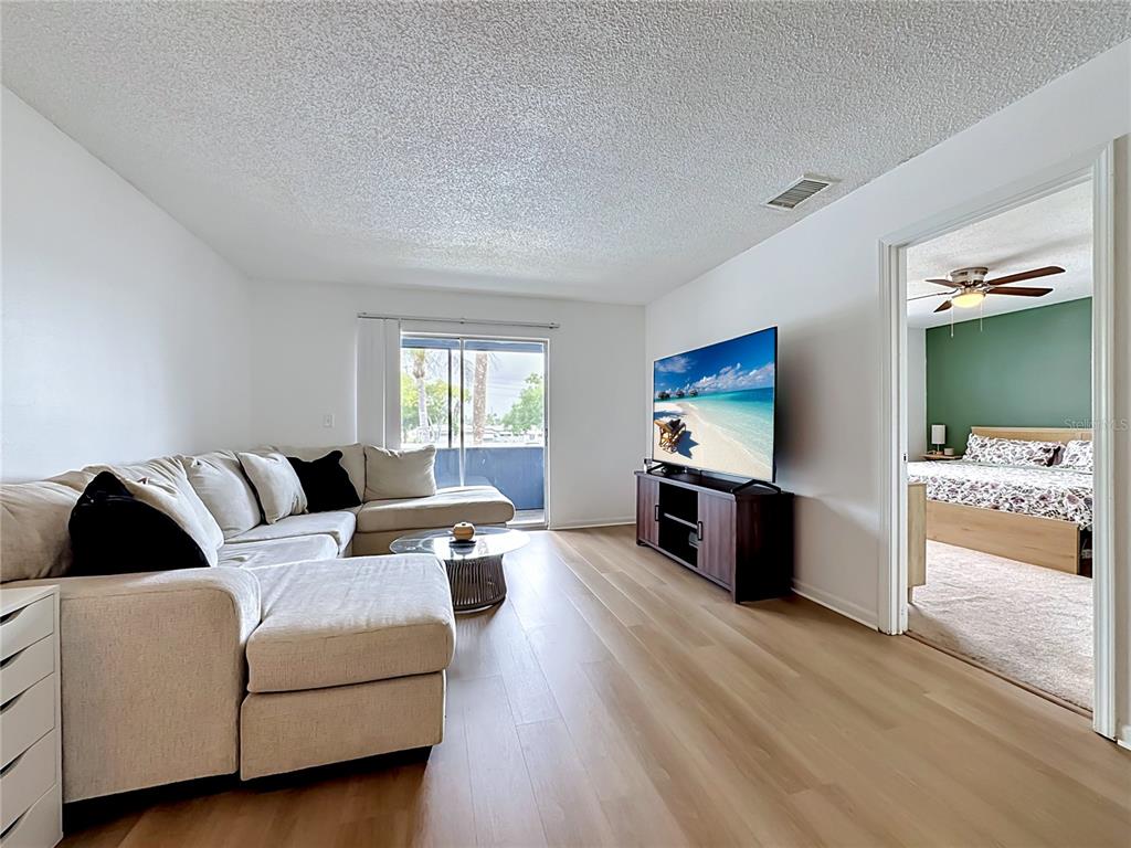 A living room with a sectional sofa, television, and wood flooring leads to a bedroom — 5507 FOUNTAIN LAKE CIRCLE UNIT B206, BRADENTON, FL
