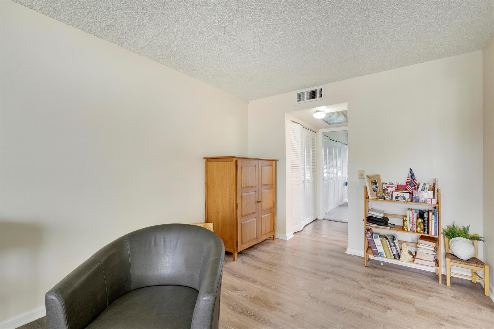 A room with light-colored walls, wood-look flooring, a gray armchair, a wooden cabinet, and shelving — 16 Mansfield A, Boca Raton FL 33434, BOCA RATON, FL