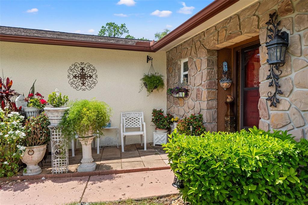 A stone-accented entryway features a patio with potted plants, a white chair, and wall decor — 10541 GREEN MEADOW LANE, PORT RICHEY, FL