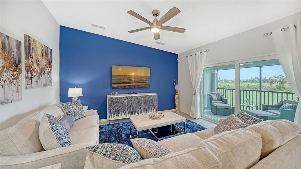 A living room features a blue accent wall, a television, and a sliding glass door — 12550 GALAPAGOS COURT UNIT 205, VENICE, FL