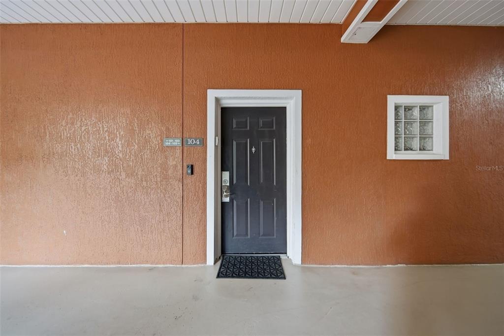 A dark front door with a glass block window sits on a textured orange wall — 9051 TREASURE TROVE LANE UNIT 104, KISSIMMEE, FL
