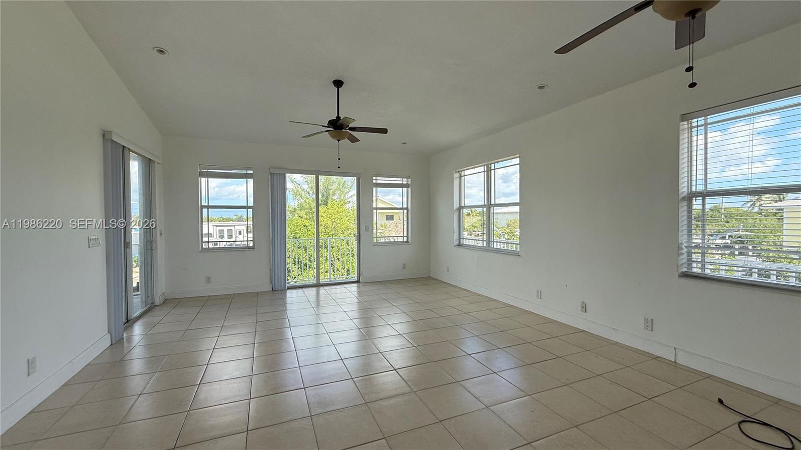 A bright, empty living room features beige tile flooring, white walls, and multiple windows — 27403 Antigua Ln, Lower Keys FL 33042, LOWER KEYS, FL