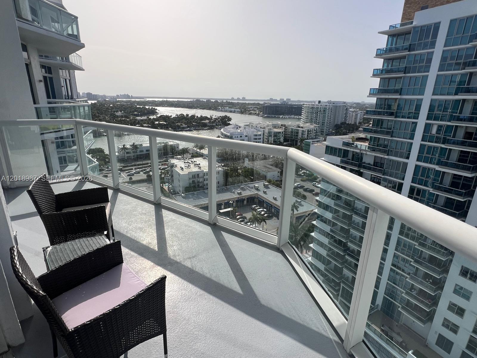 A high-rise balcony with wicker chairs overlooks a city waterway and surrounding buildings — 6515 Collins Ave # 1801, Miami Beach FL 33141, MIAMI BEACH, FL