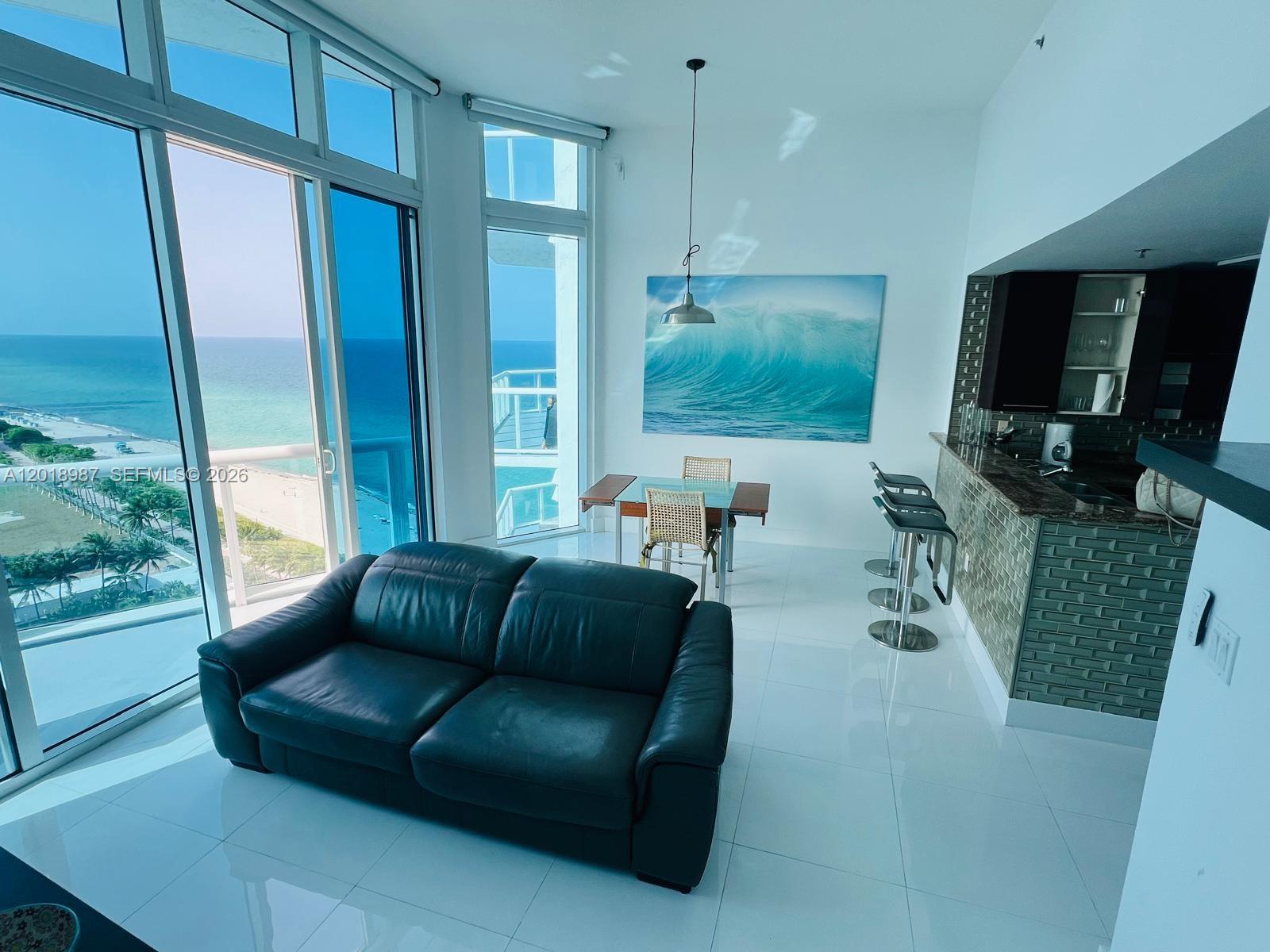 A modern living area features a dark leather sofa, dining table, and oceanfront balcony views — 6515 Collins Ave # 1801, Miami Beach FL 33141, MIAMI BEACH, FL