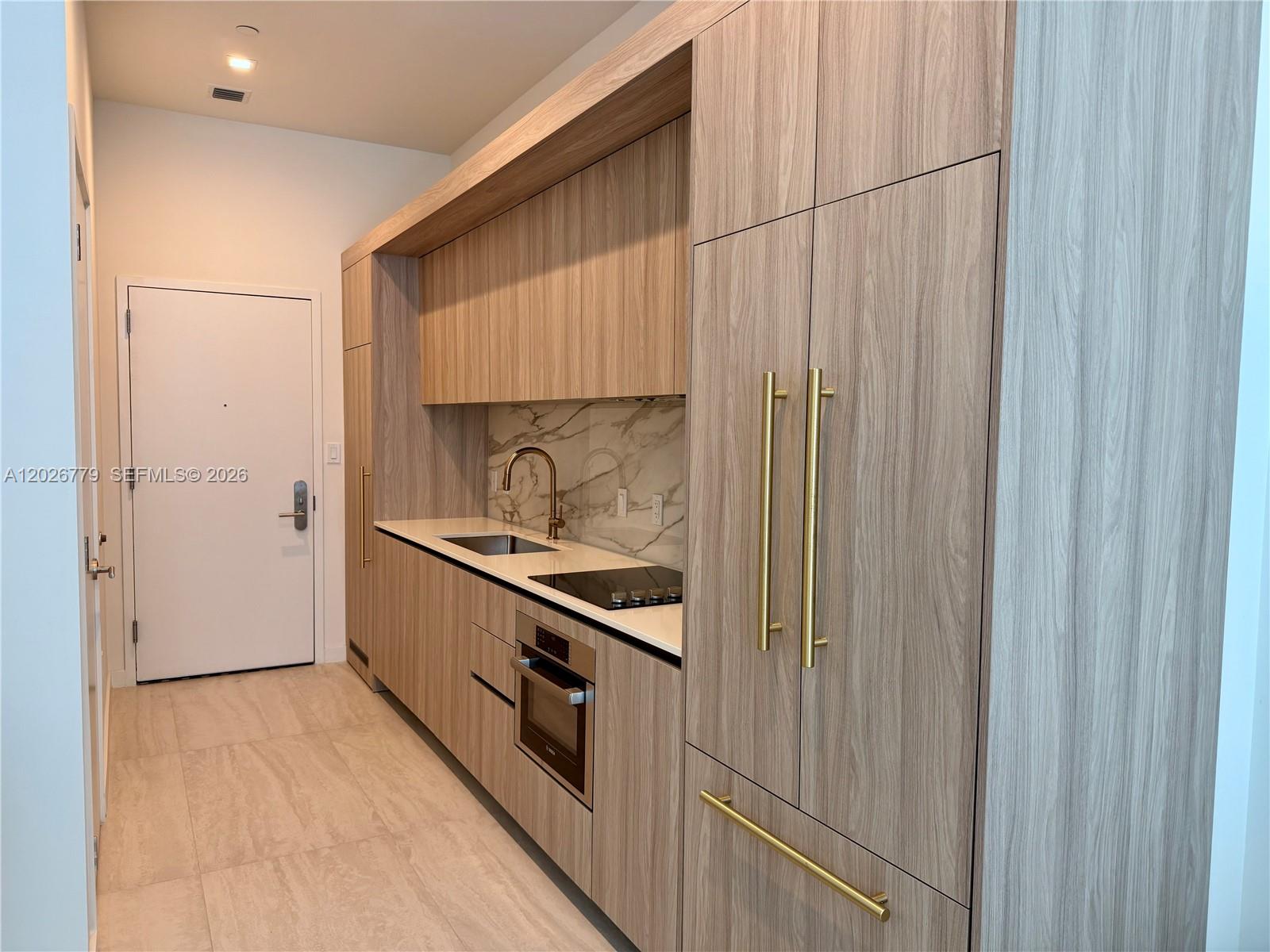 A modern kitchenette features wood-grain cabinetry, gold hardware, a marble backsplash, and integrated appliances — 77 SE 5th St # 407, Miami FL 33131, MIAMI, FL