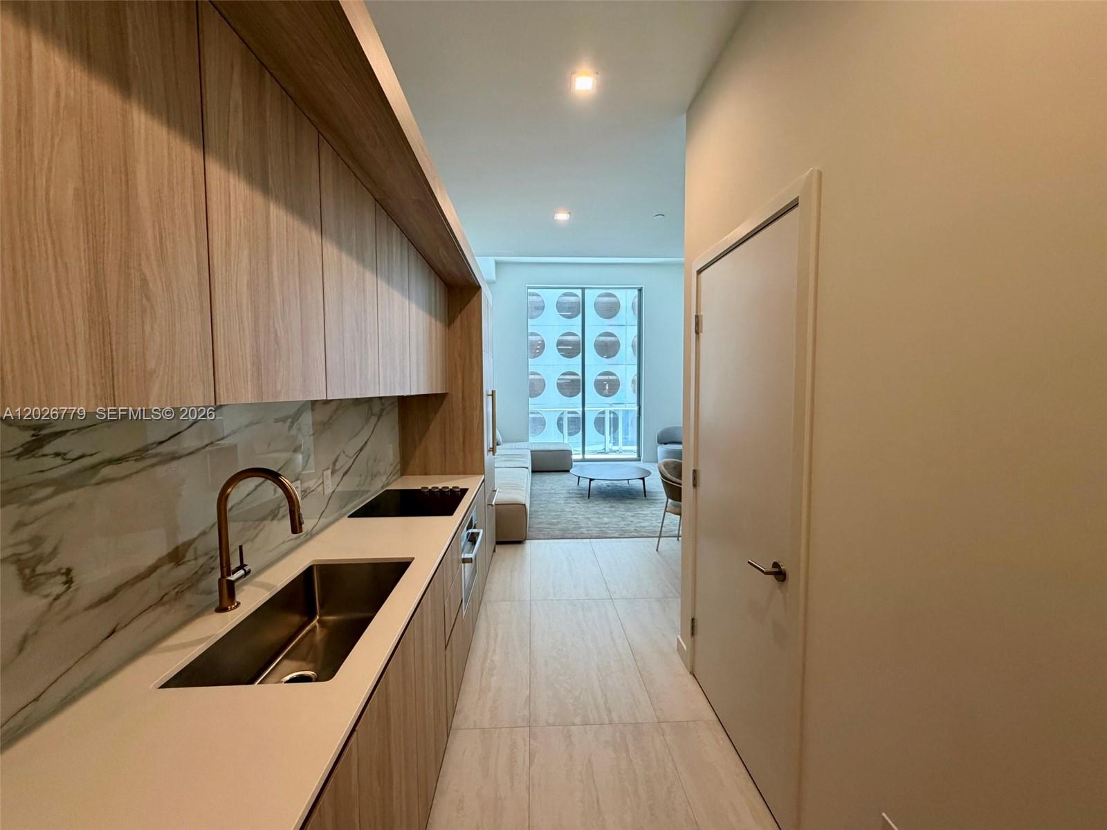 A modern kitchen with wood cabinetry and marble backsplash leads to a living area — 77 SE 5th St # 407, Miami FL 33131, MIAMI, FL