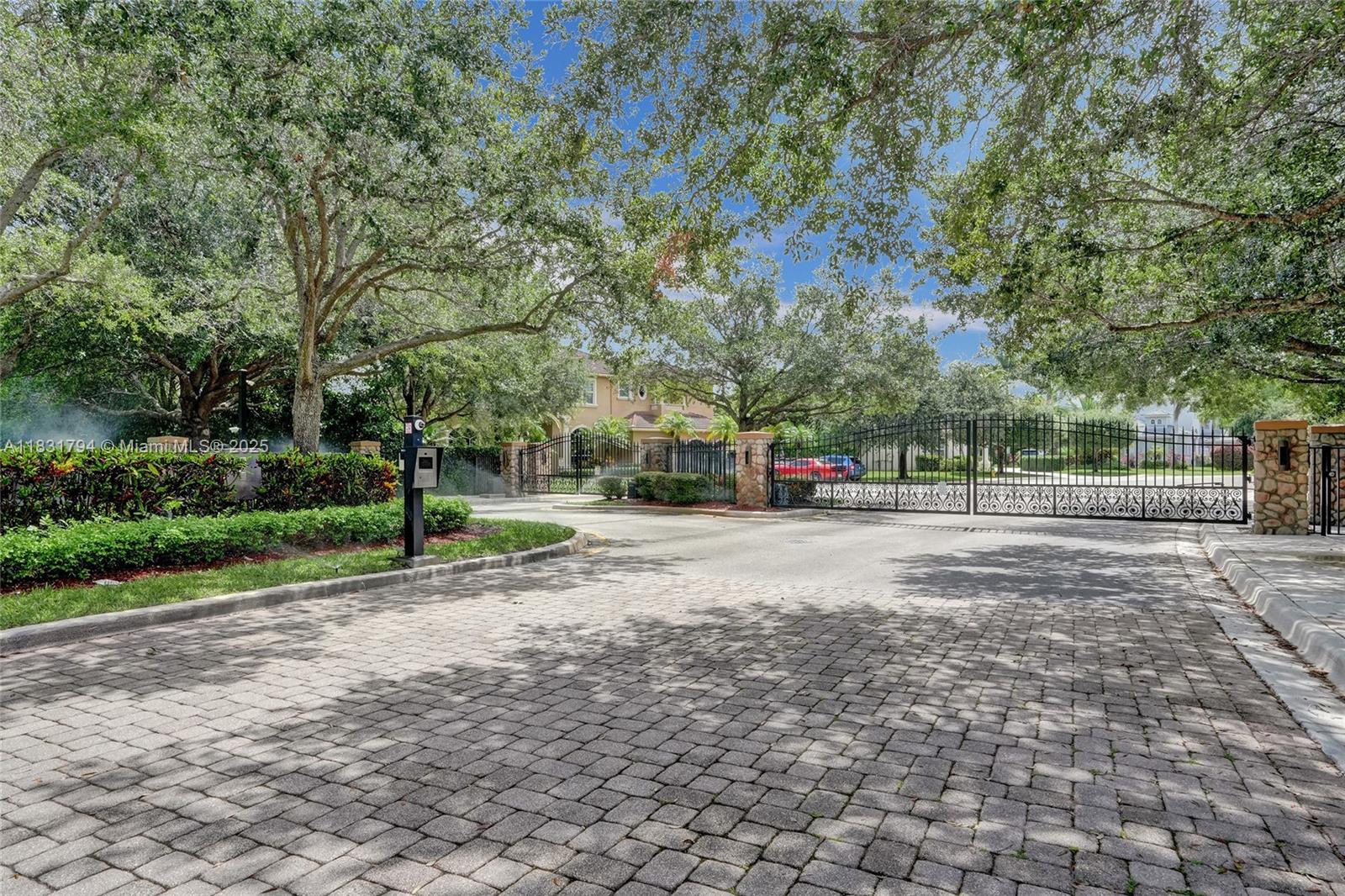 A gated entrance with a paved driveway leads to a residential community under large trees — 11056 Blackhawk Blvd, Davie FL 33328, DAVIE, FL