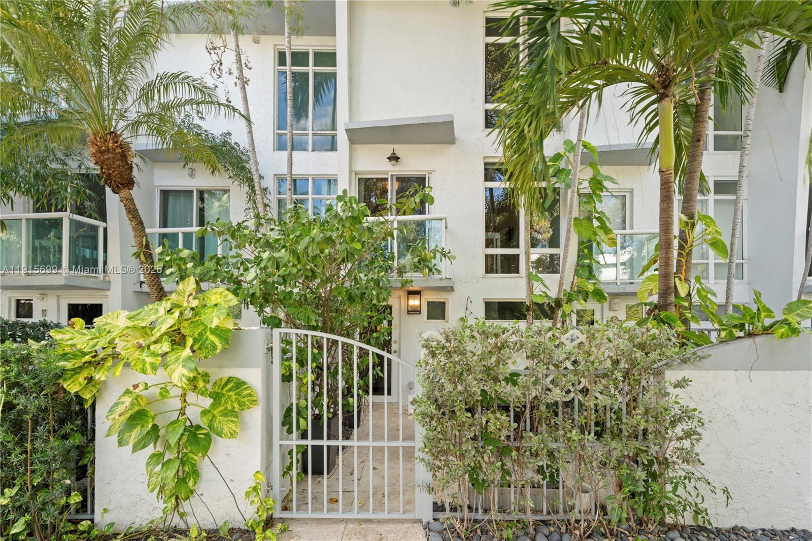 A white multi-story townhouse exterior features a gated entrance, balconies, and lush tropical landscaping — 1853 Jefferson Ave # 6, Miami Beach FL 33139, MIAMI BEACH, FL