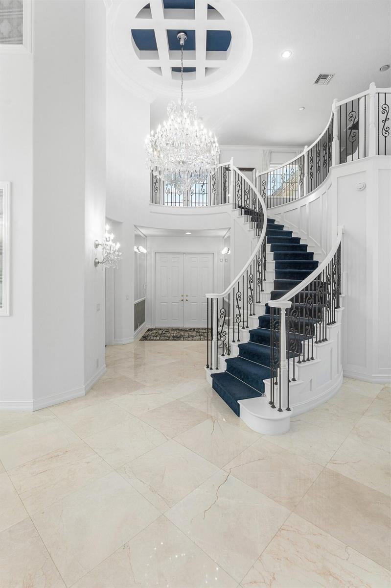 A grand foyer features a curved staircase with blue carpet, marble flooring, and a chandelier — 131 Carmela Court, Jupiter FL 33478, JUPITER, FL