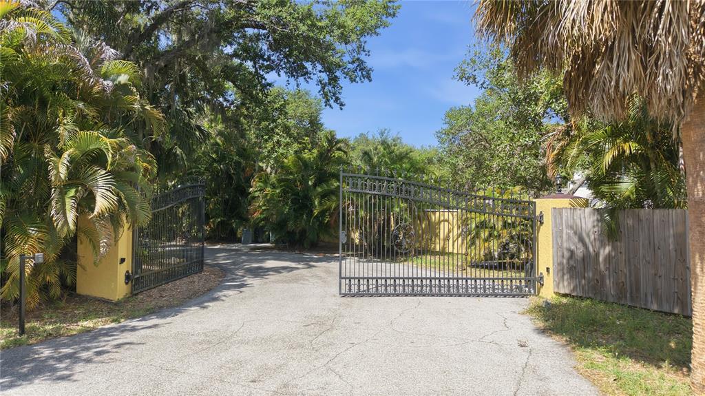 An open wrought iron gate leads to a paved driveway surrounded by lush tropical landscaping — 3348 ALAMO LANE, SARASOTA, FL