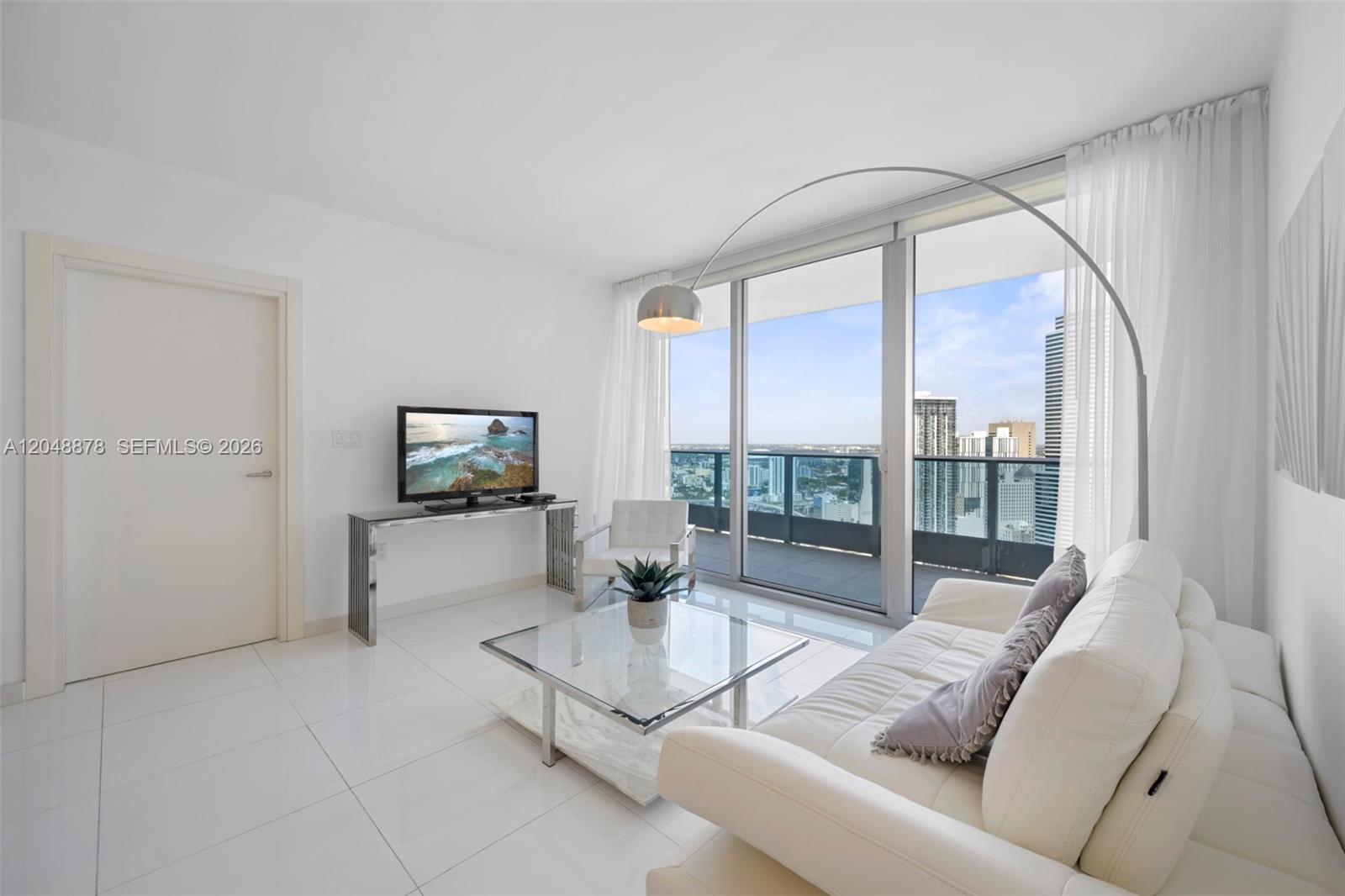 A modern living room features white tile floors, a glass coffee table, and a balcony view — 200 Biscayne Boulevard Way # 4314, Miami FL 33131, MIAMI, FL