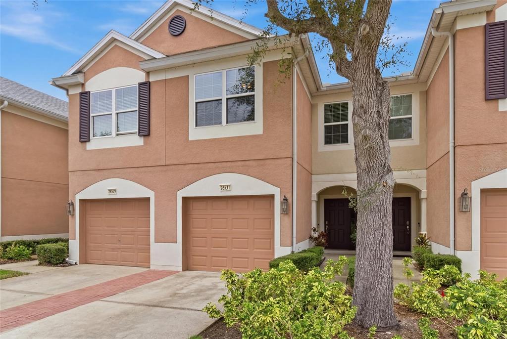 A two-story townhouse exterior features tan stucco walls, two garage doors, and a front entrance — 26531 CHIMNEY SPIRE LANE, WESLEY CHAPEL, FL