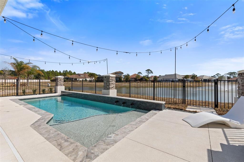 A backyard swimming pool with a water feature overlooks a lake behind a metal fence — 122 DEL PALMA DRIVE, PALM COAST, FL