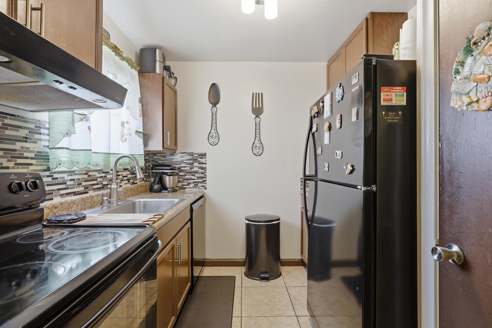 A galley-style kitchen features wood cabinets, black appliances, a tiled backsplash, and a tile floor — 6074 Forest Hill Boulevard # 101, West Palm Beach FL 33415, WEST PALM BEACH, FL