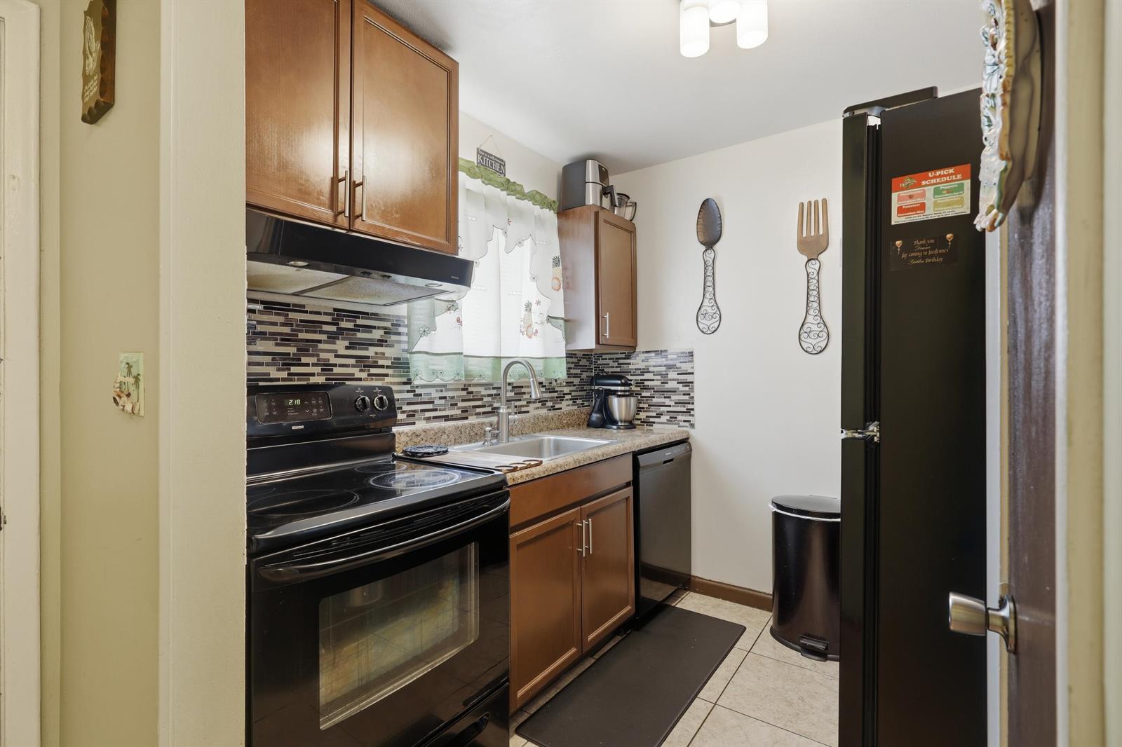 A galley kitchen features wood cabinets, black appliances, a tiled backsplash, and light-colored floor tiles — 6074 Forest Hill Boulevard # 101, West Palm Beach FL 33415, WEST PALM BEACH, FL