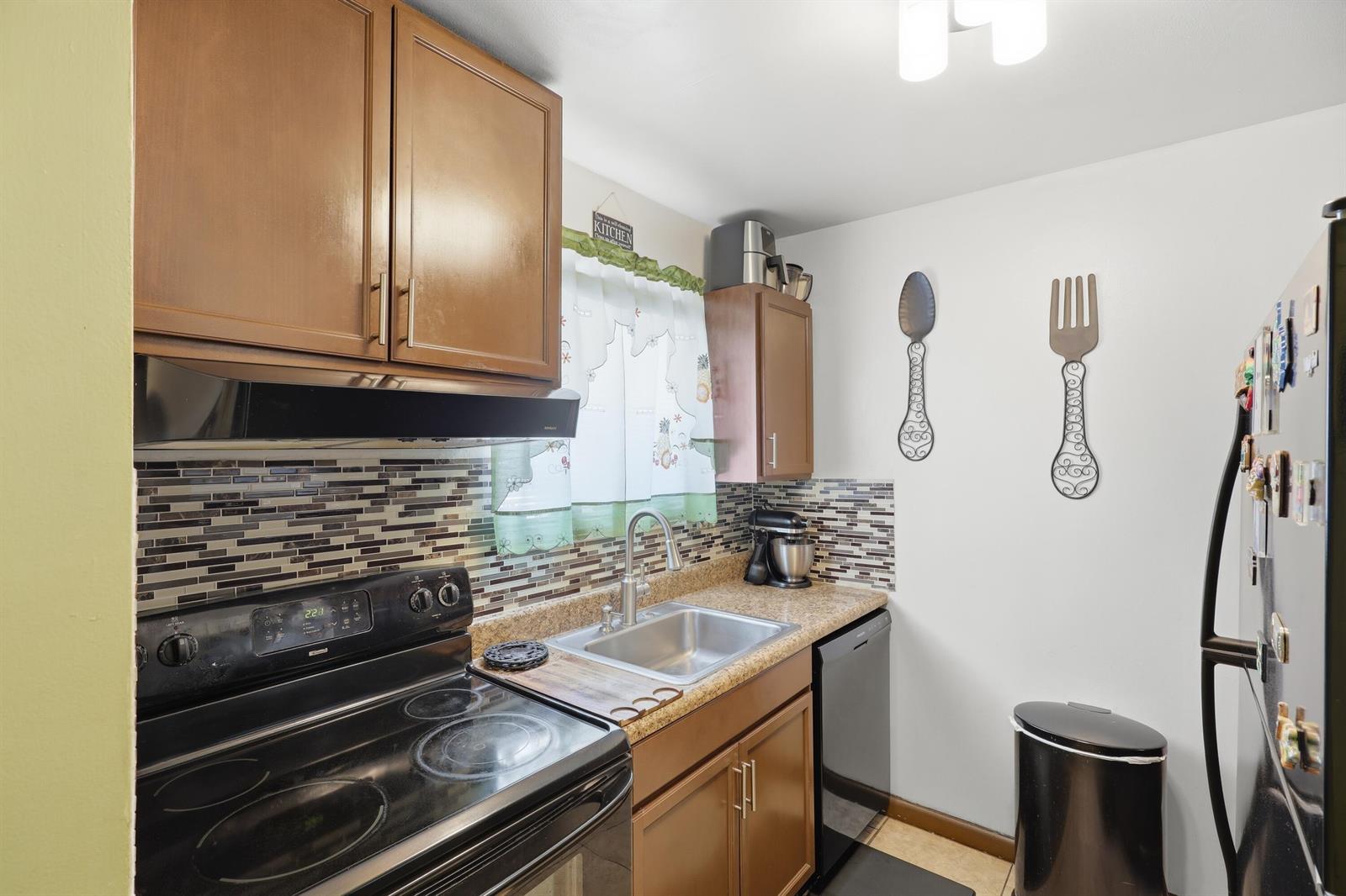 A small kitchen features brown cabinets, a black stove, a stainless steel sink, and tile backsplash — 6074 Forest Hill Boulevard # 101, West Palm Beach FL 33415, WEST PALM BEACH, FL
