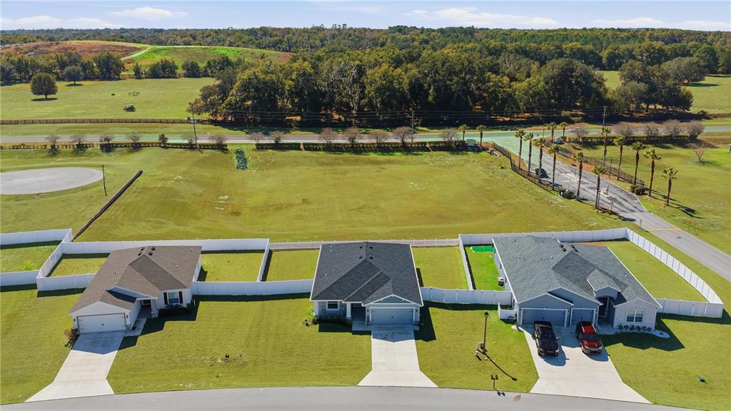 An aerial view shows three suburban houses with white fences bordering a large, grassy field — 4810 SE 91ST PLACE, OCALA, FL