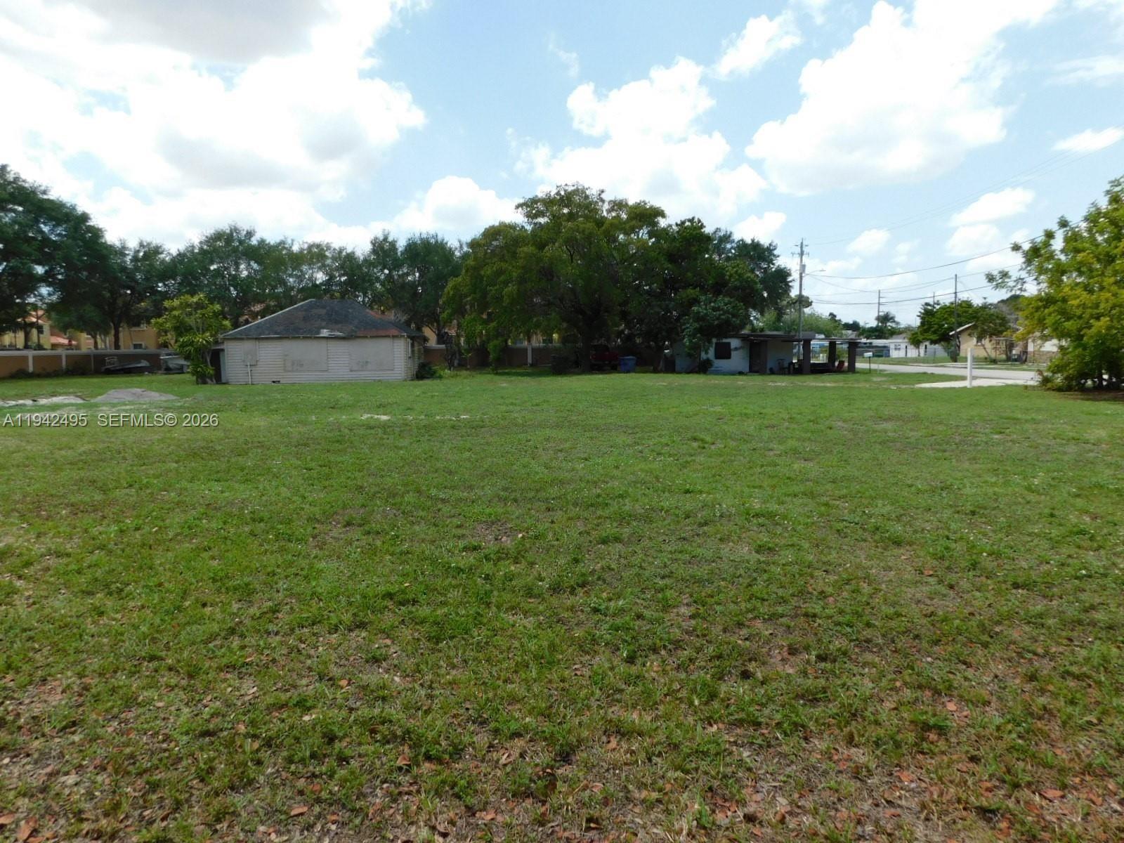 A grassy vacant lot features a small, weathered building and mature trees in the background — 5830 Liberty St, Hollywood FL 33021, HOLLYWOOD, FL