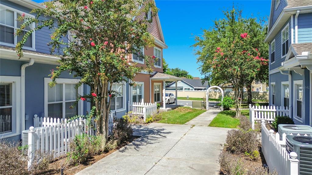 A concrete walkway leads between two blue townhouses with white picket fences and flowering trees — 2208 SAN VITTORINO CIRCLE UNIT 104, KISSIMMEE, FL