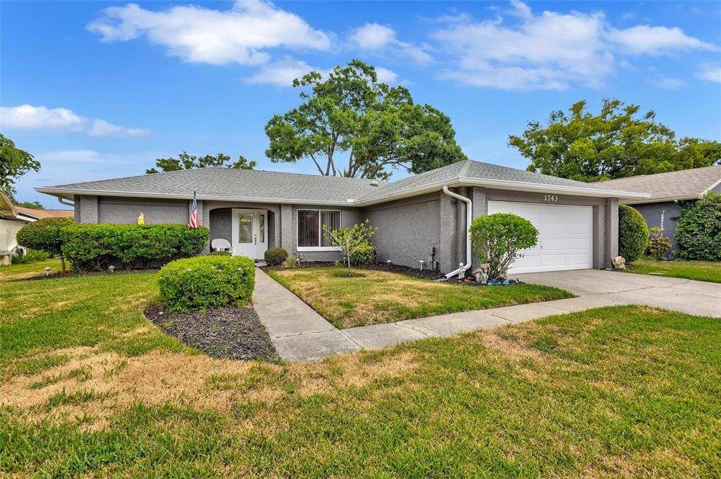 A single-story gray house with a white garage door, front walkway, and green landscaping — 1743 MACDONNELL DRIVE, PALM HARBOR, FL