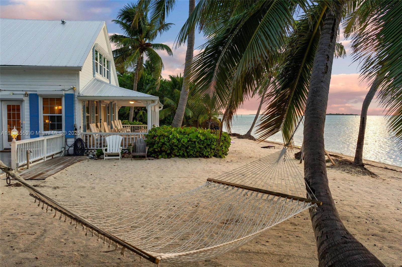 A white house with blue shutters sits on a sandy beach beside a hammock — 109 Columbus Dr, Islamorada FL 33036, ISLAMORADA, FL