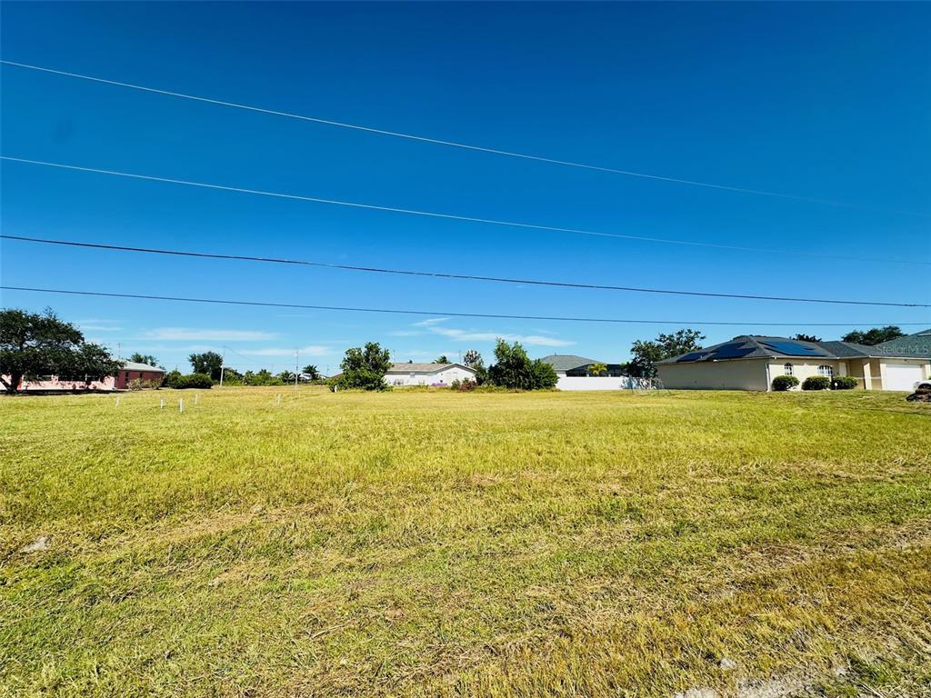 A wide, grassy vacant lot sits under a clear blue sky with neighboring houses visible — 2053 NW 3RD STREET, CAPE CORAL, FL