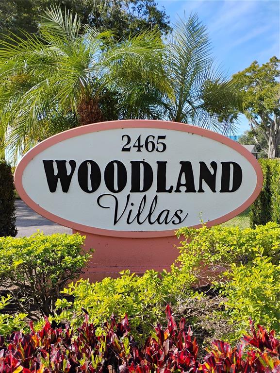 An oval sign for Woodland Villas stands in front of a palm tree and bushes — 2465 NORTHSIDE DRIVE UNIT 1001, CLEARWATER, FL