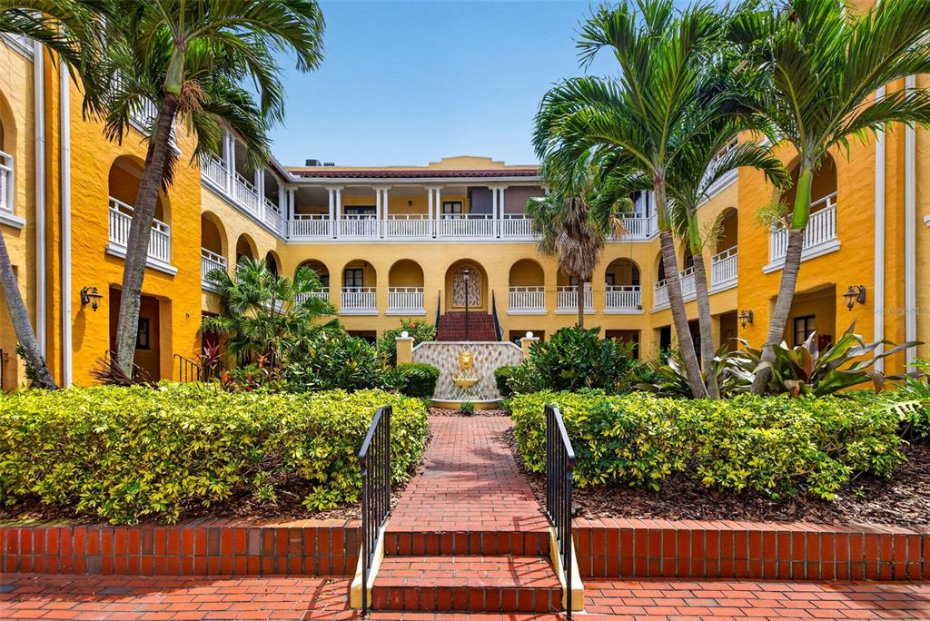 A courtyard with a brick path, fountain, and palm trees surrounded by yellow multi-story buildings — 210 22ND AVENUE NE UNIT 12, ST PETERSBURG, FL
