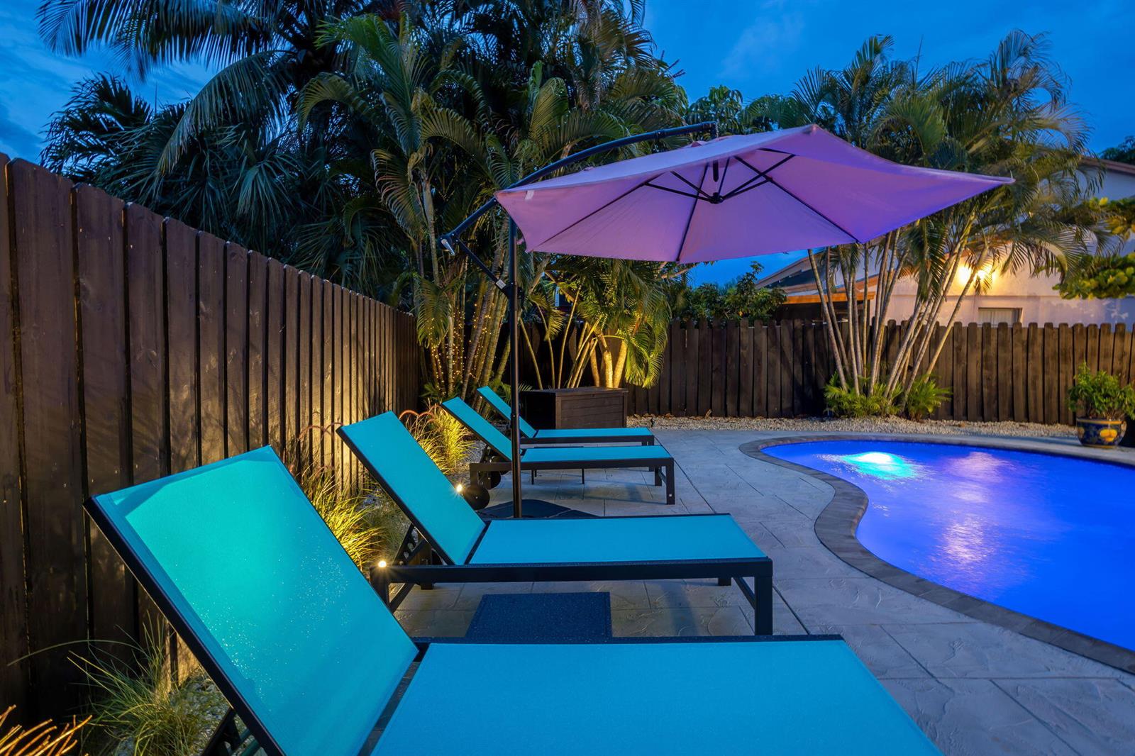 A backyard pool area features three turquoise lounge chairs under a purple umbrella at dusk — 2541 Southridge Road, Delray Beach FL 33444, DELRAY BEACH, FL