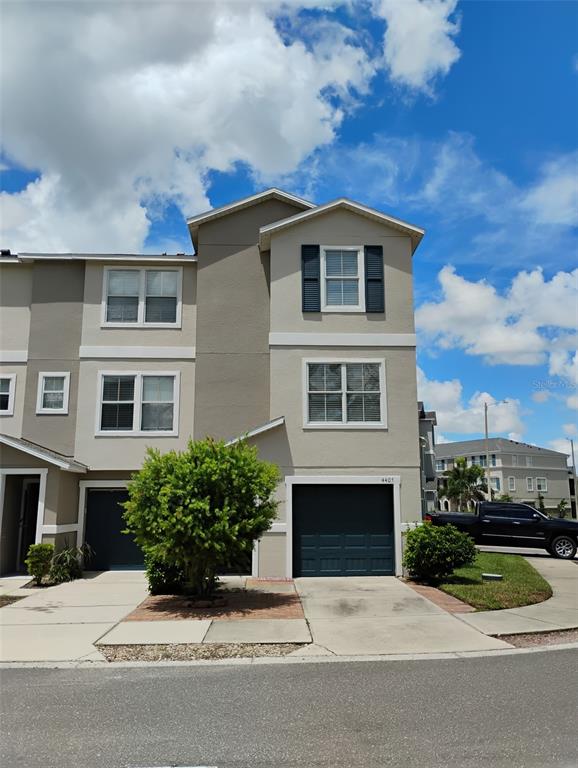 A three-story townhouse exterior features a dark garage door, beige siding, and a small tree — 4405 TUSCAN LOON DRIVE, TAMPA, FL