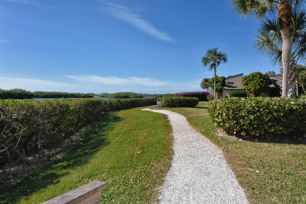 A gravel path winds through a grassy lawn toward a coastal landscape with palm trees — 1712 GLENHOUSE DRIVE UNIT GL418, SARASOTA, FL