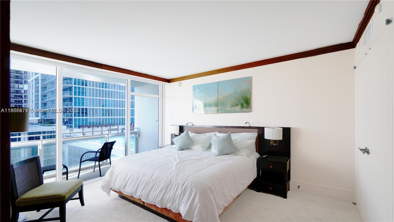 A bedroom features a bed with light blue pillows, dark nightstands, and a balcony view — 6801 Collins Ave # 710, Miami Beach FL 33141, MIAMI BEACH, FL