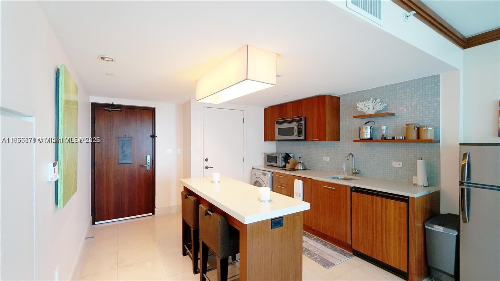 A kitchen features wooden cabinets, a white island with stools, and light-colored tile flooring — 6801 Collins Ave # 710, Miami Beach FL 33141, MIAMI BEACH, FL