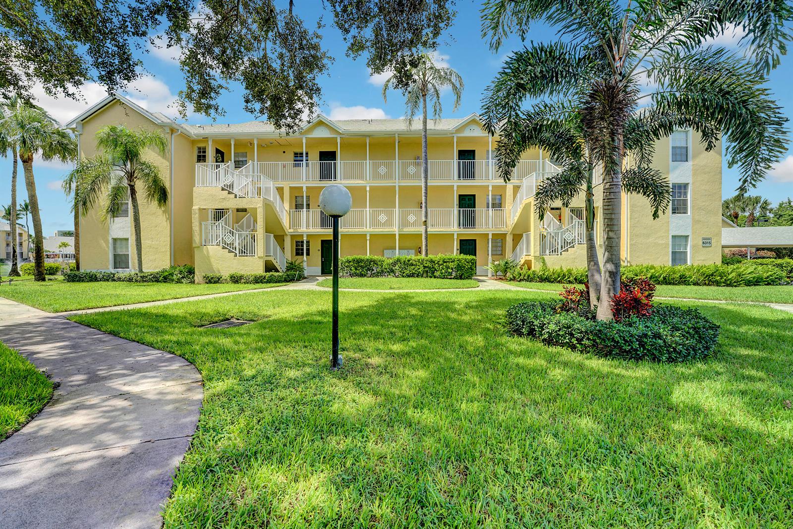 A three-story yellow apartment building features exterior staircases, white railings, and a landscaped lawn — 6315 La Costa Drive # A, Boca Raton FL 33433, BOCA RATON, FL