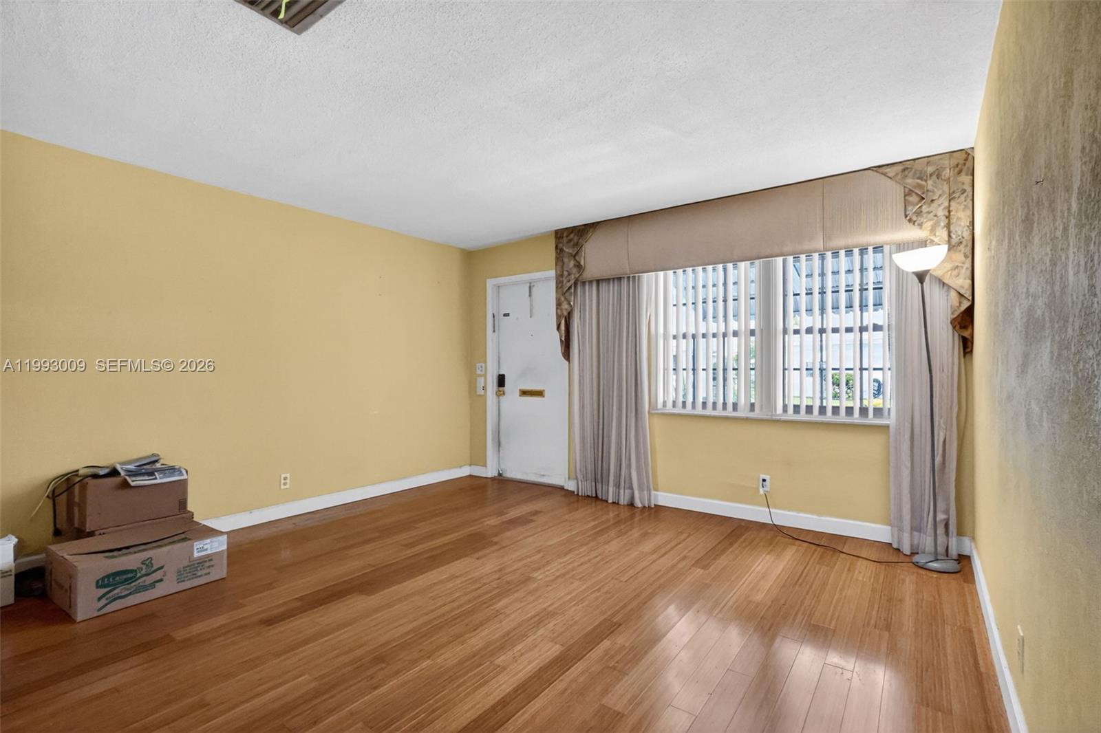 An empty room features yellow walls, hardwood flooring, a window with curtains, and a door — 1104 Ocean Ave, Boynton Beach FL 33426, BOYNTON BEACH, FL